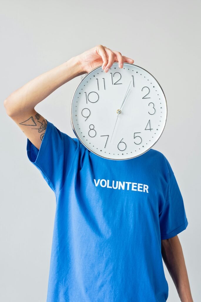 A person in a blue volunteer shirt covering their face with a large clock.